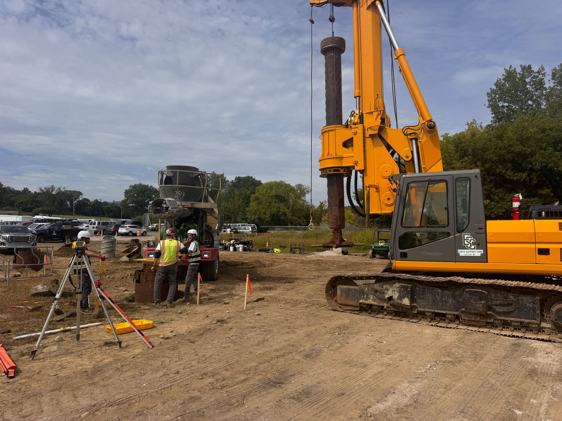 An orange drill rig with its kelly bar raised over a hole on a gravel lot, with crew in safety vests and a survey instrument on a tripod in the foreground.