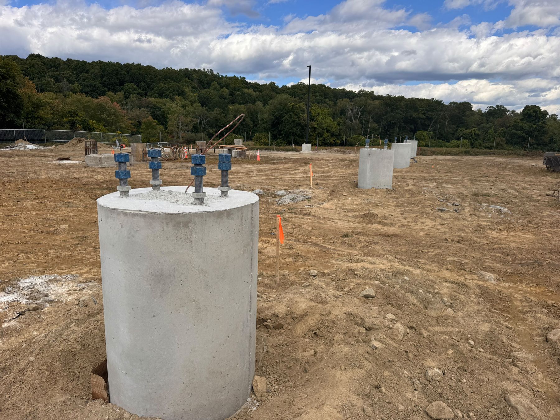 A round concrete pier standing out of the ground with four threaded anchor rods capped in blue, on a graded site with more piers in a row behind and a wooded hillside beyond.