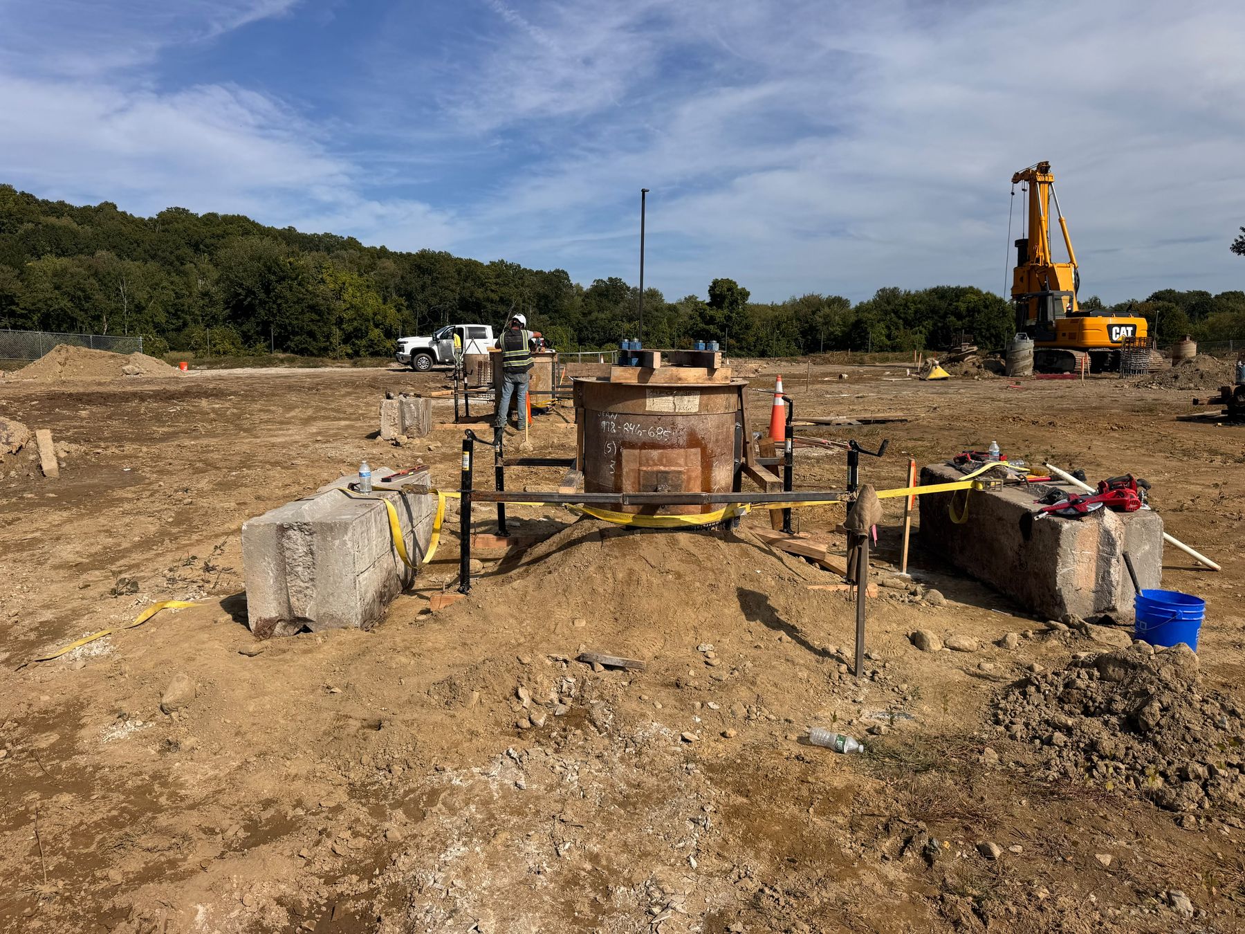 A round steel form over a drilled hole, held by a frame on adjustable stands between two concrete blocks, with a worker beside it and the drill rig on the next hole behind.