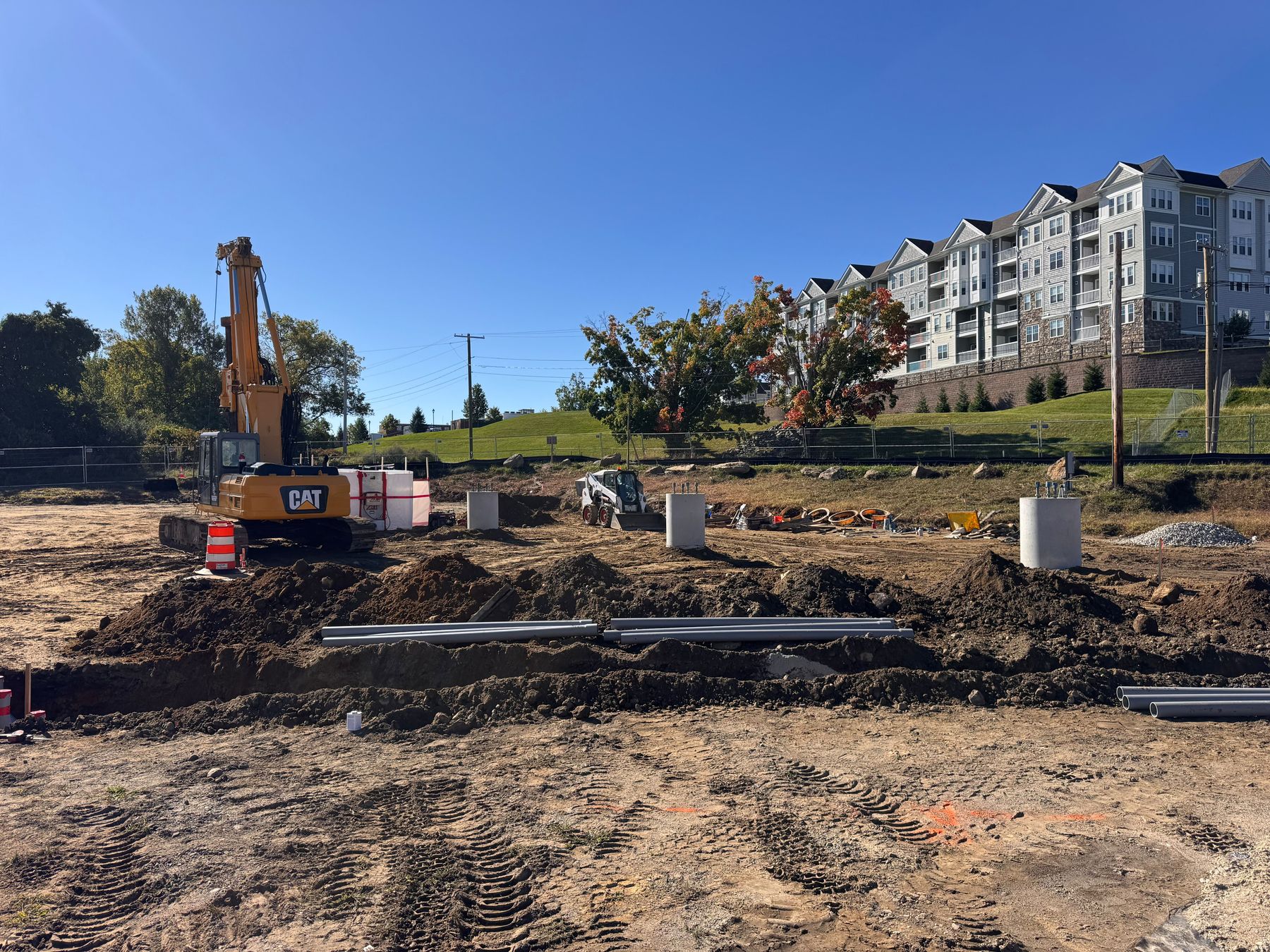 A graded construction site with several finished concrete piers, the drill rig, a compact loader, and conduit pipe beside a trench, with an apartment building on the hill behind.
