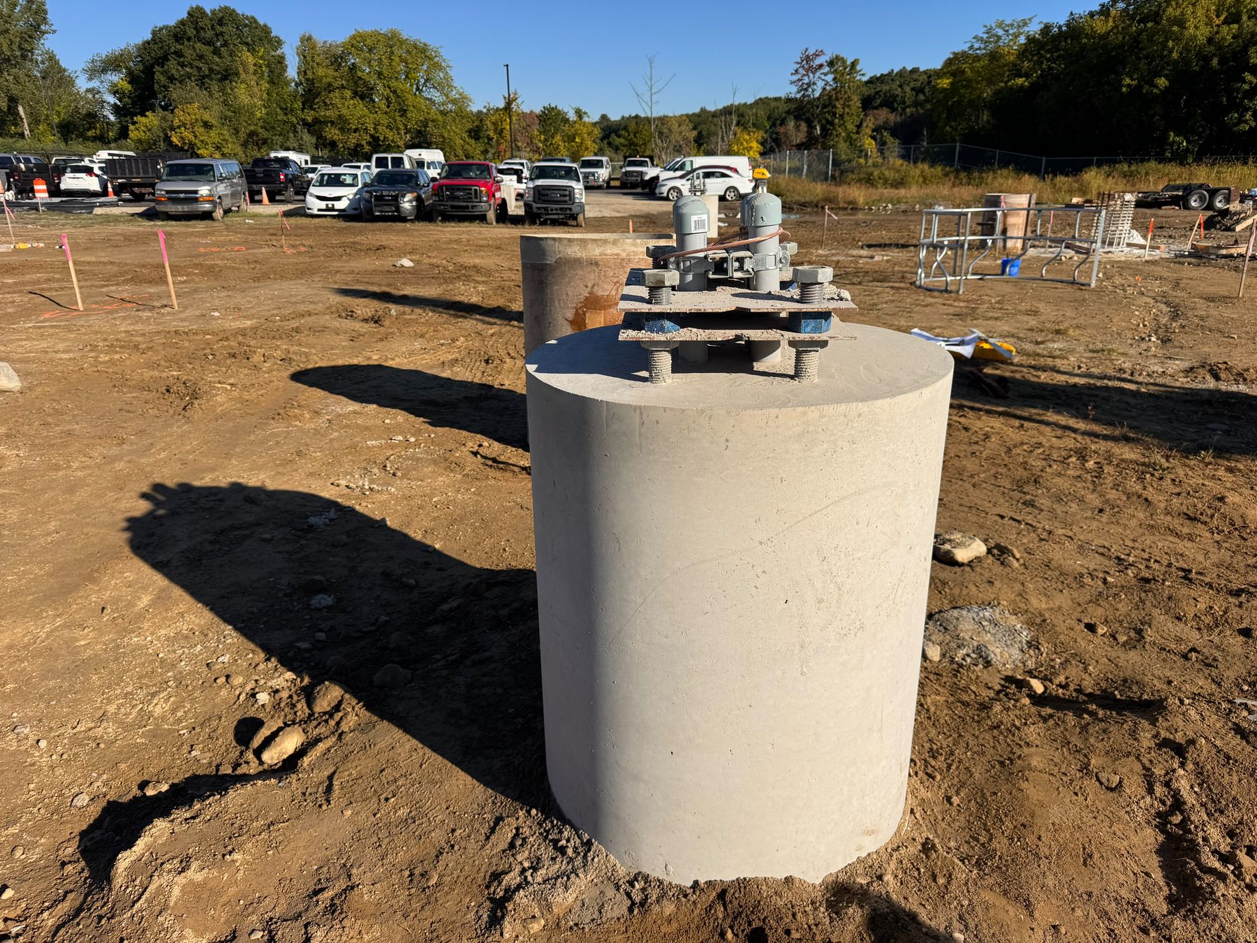 A freshly poured round concrete pier with a steel anchor-rod template still bolted on top, beside a parking lot full of vehicles.