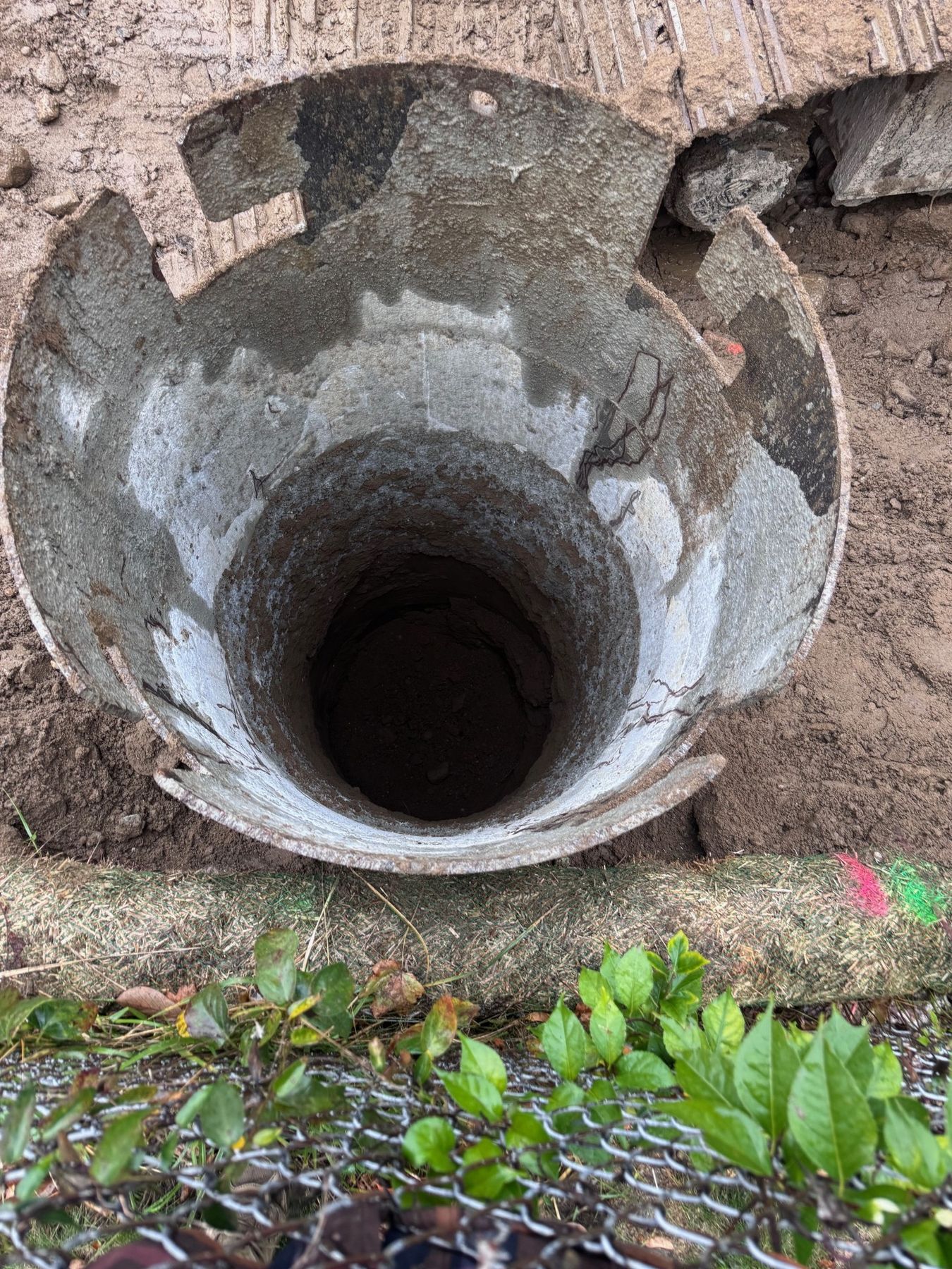 Looking down into a round drilled hole lined with temporary steel casing, beside the property-line fence.