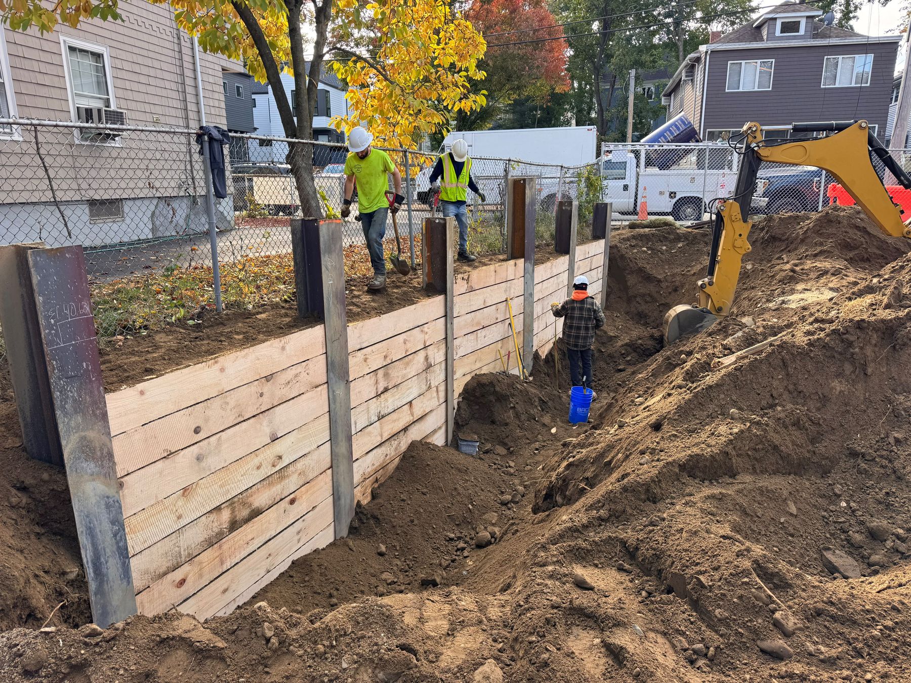 A timber-lagged soldier pile wall along a chain-link fence, with two workers in hard hats and vests at the top, one in the cut, and an excavator digging at the right, with houses behind.