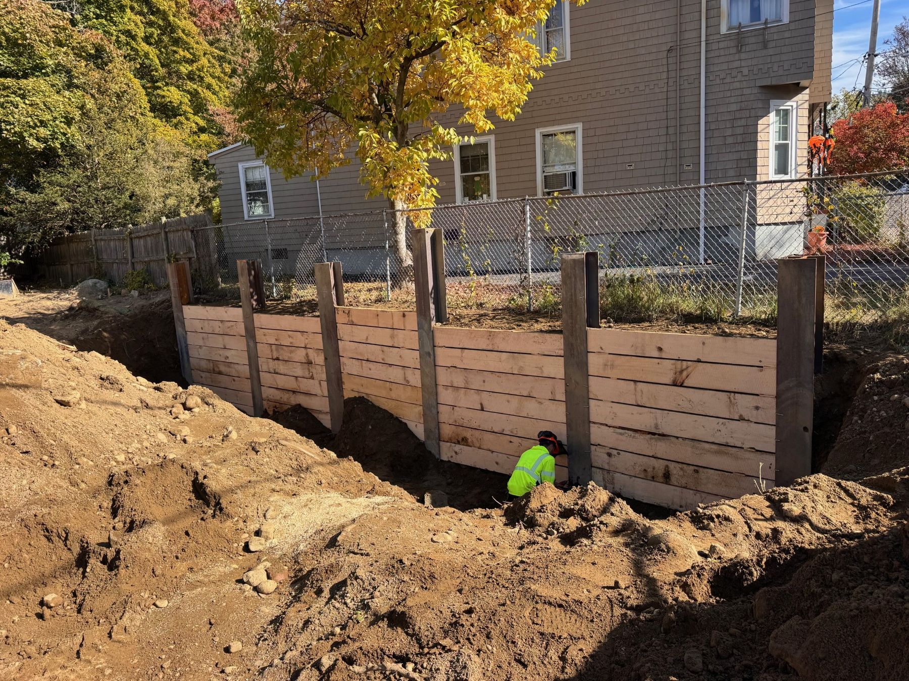 An excavation in front of a timber-lagged soldier pile wall, with the neighbor's chain-link fence and house directly behind it.