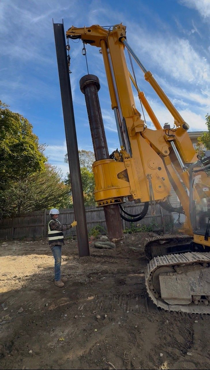 The drill rig lowering a steel H-pile toward a drilled hole as a crew member guides it.
