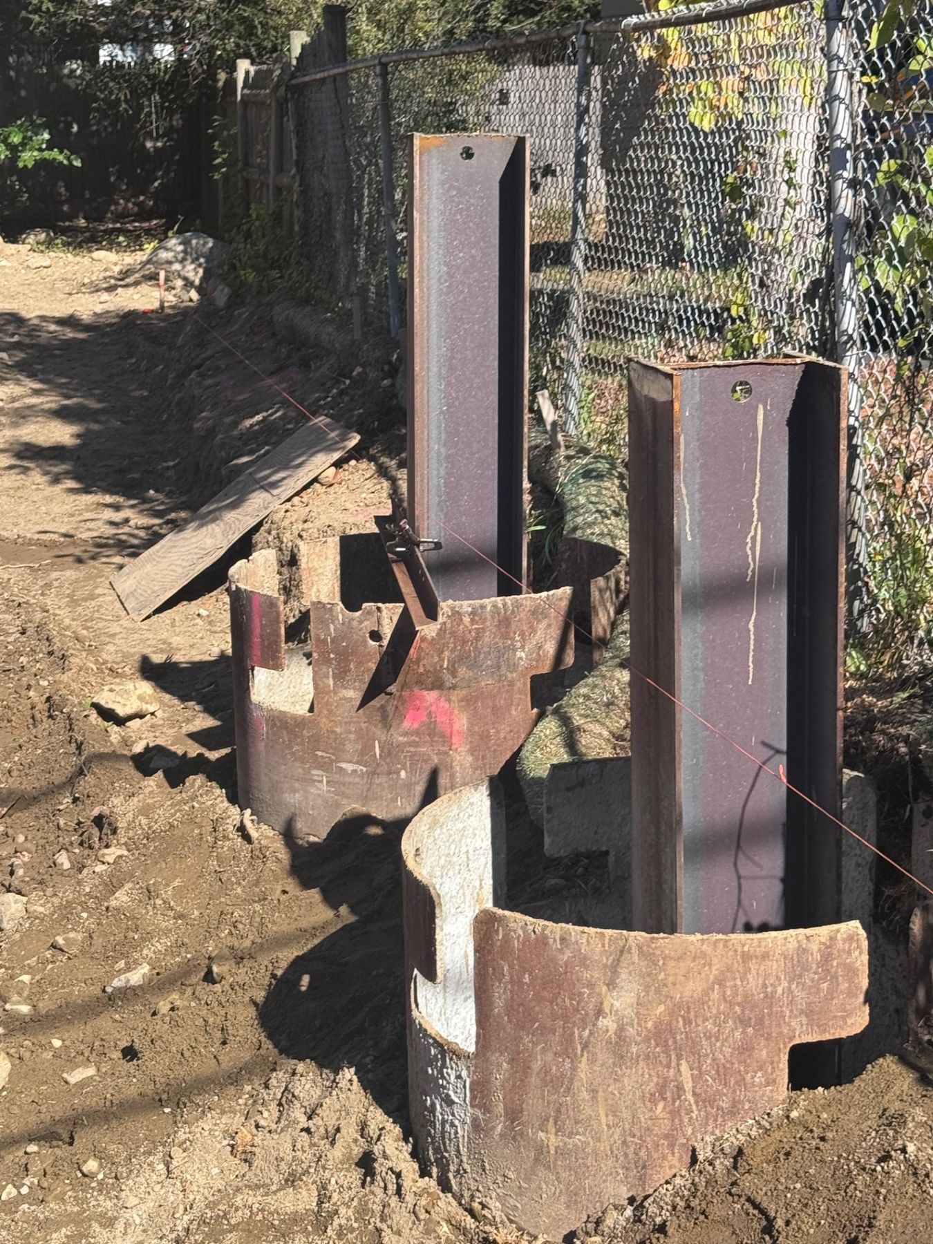 Two steel H-piles standing in backfilled holes next to a chain-link fence, with cut sections of steel casing at their base.