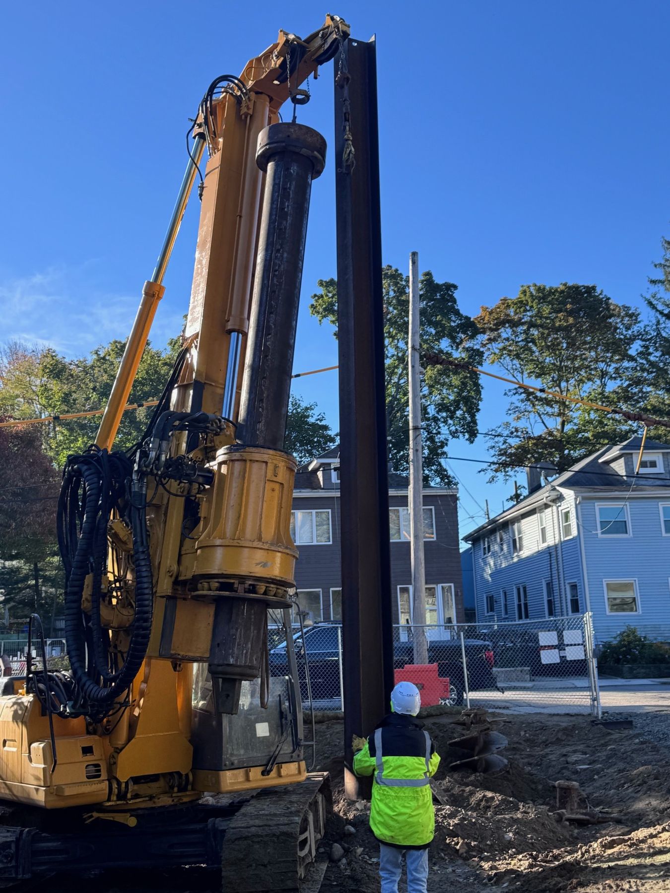 A drill rig lifting a steel H-pile upright on a residential lot while a worker in a hard hat and safety vest looks on.